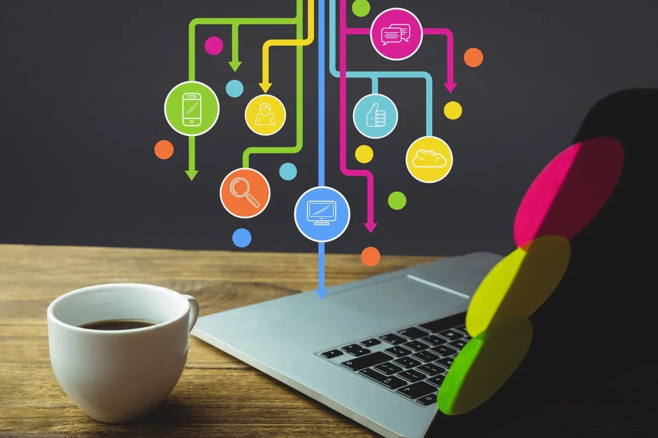 Cup of coffee next to a laptop with colorful digital tool icons floating above it on a wooden desk.
