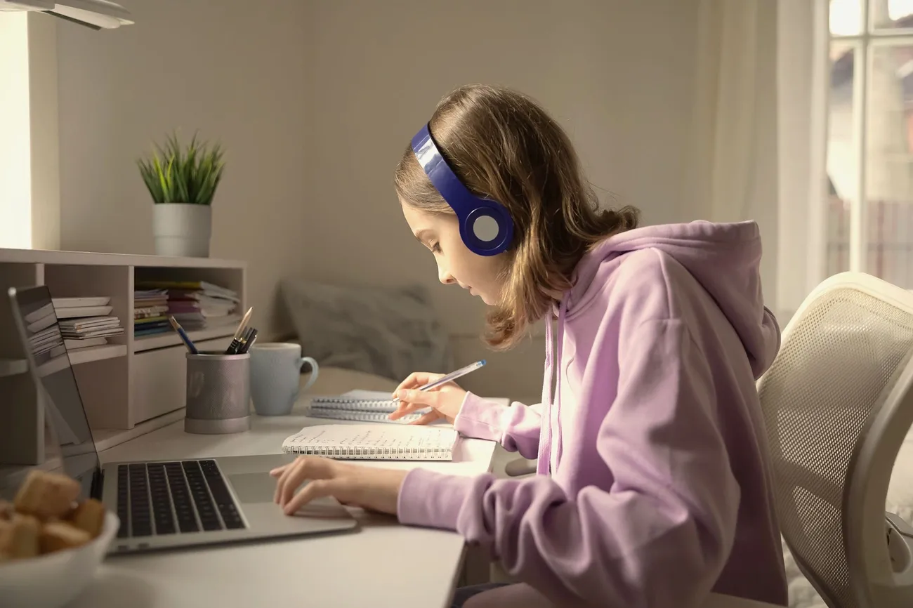 Student in a pink hoodie studies at a desk with a laptop, notebook, and pencil holder in a bright room.