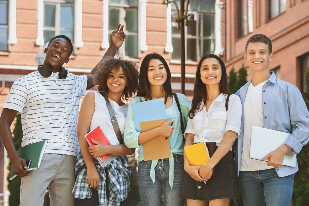 Group of five diverse college students standing outside campus buildings holding notebooks and a laptop during orientation.