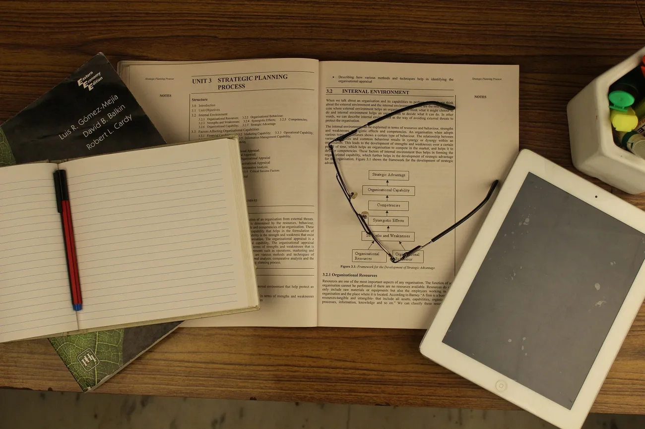 A study setup showing open paper notes, a pen, reading glasses, and a tablet on a wooden desk symbolizing digital vs. paper notes.