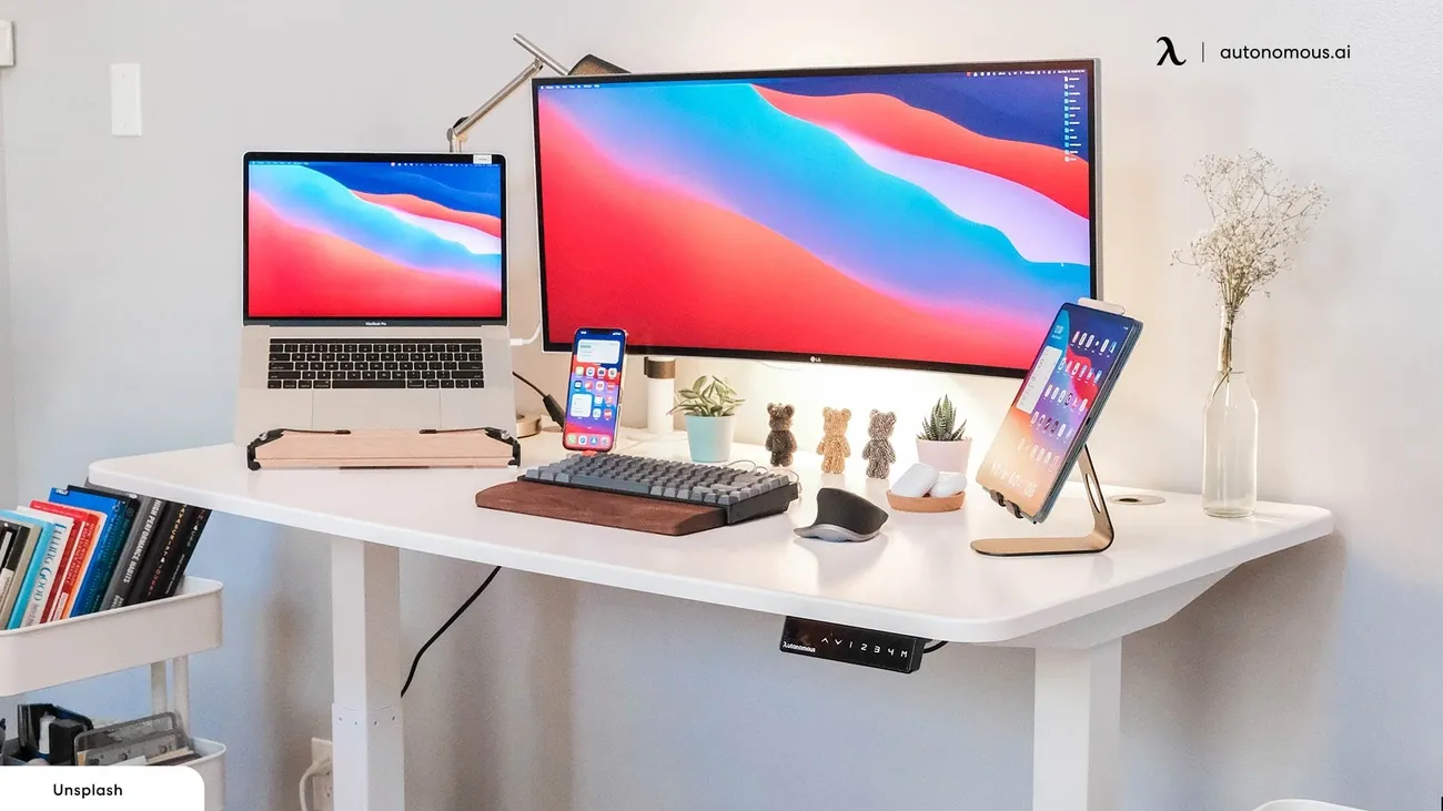 Minimalist college desk setup with laptop, ultra-wide monitor, tablet, smartphone, keyboard, mouse, and decorative plants and figurines.