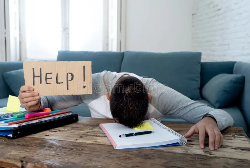 Frustrated student with head down on notebook holding a cardboard sign that says 'HELP!' at a wooden table.