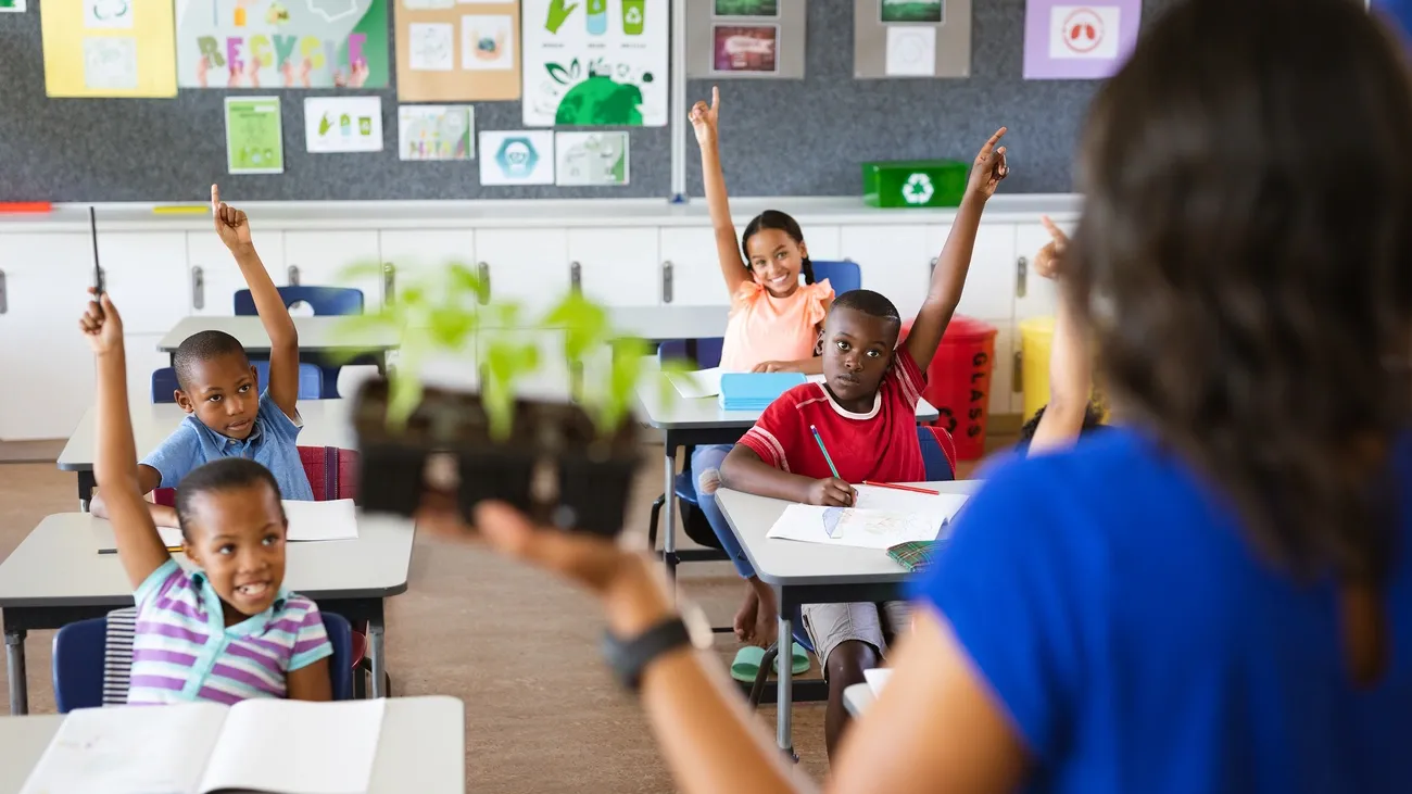 Teacher holds seedlings while students in a classroom eagerly raise their hands during a lesson on climate change.
