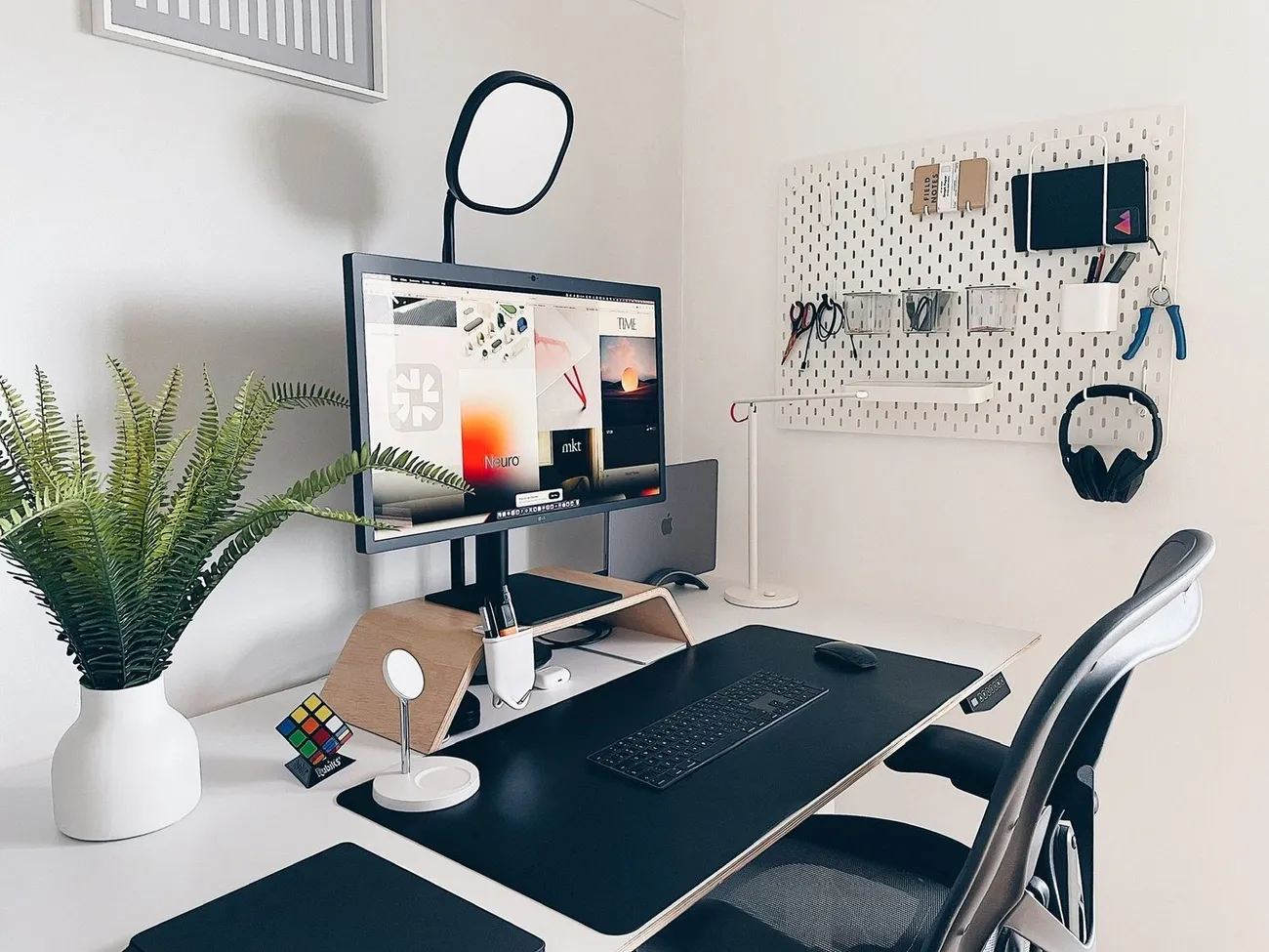 Minimalist desk setup with a monitor on a wooden stand, keyboard, mouse, plant, pegboard organizer, and ergonomic chair in a bright room.