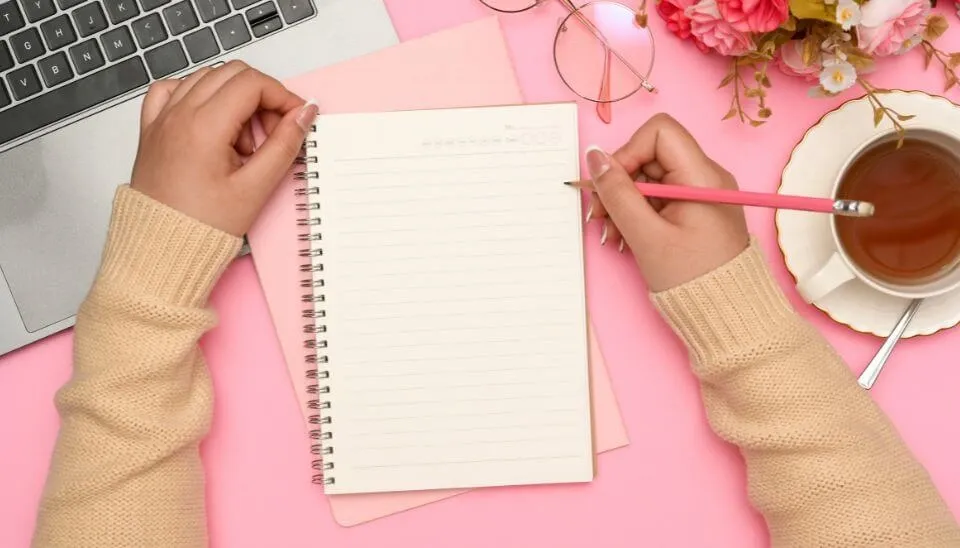 Person holding a pencil over a blank notebook on a pink desk with a laptop, glasses, flowers, and a cup of tea