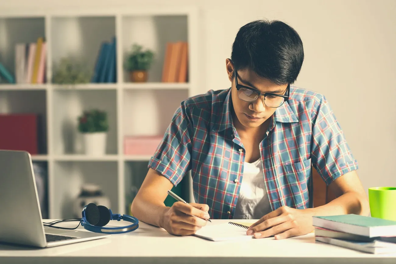 Student in a plaid shirt writing in a notebook at a desk with a laptop, headphones, and books nearby.