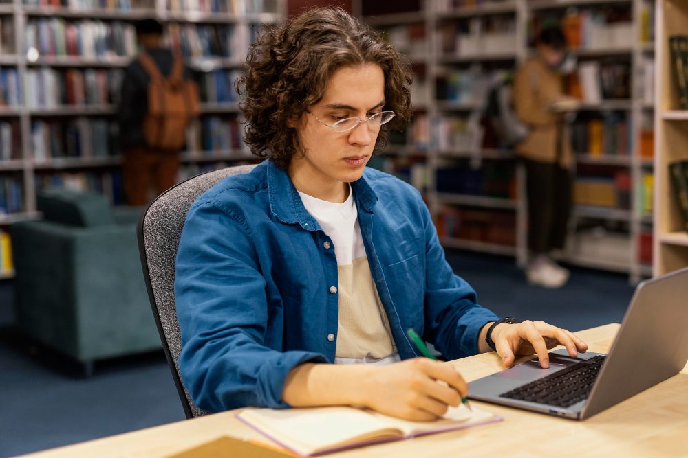 male student sitting in library infront of laptop wearing a blue jeans jacket