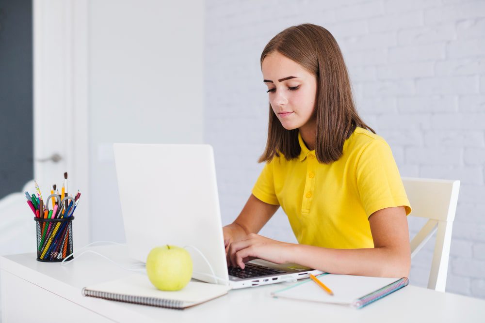 female student typing on her white laptop wearing a yellow tshirt